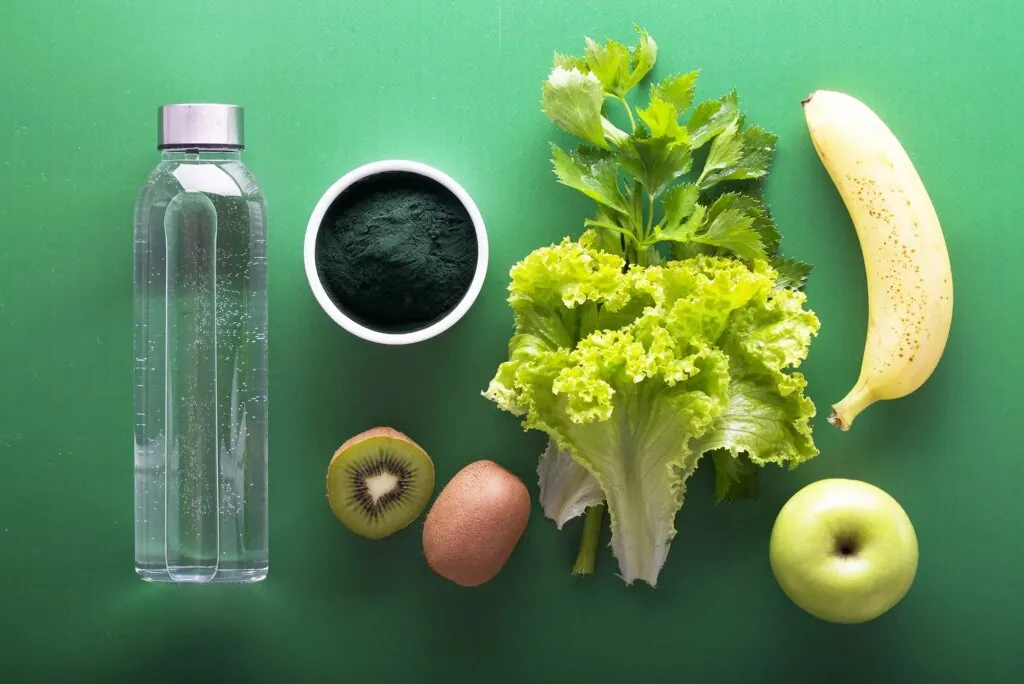 Sticlă cu apă, spirulină, salată verde, kiwi, măr și banană pe fundal verde